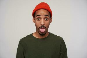 Close-up of young attractive bearded dark skinned man raising surprisedly eyebrows and showing his tongue to camera, making fun while posing over white background photo