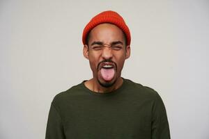 Indoor shot of young pretty bearded dark skinned guy frowning his face and showing tongue while expressing disgust, standing against white background in casual wear photo