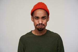 Portrait of young brown-eyed bearded dark skinned male taking air into his mouth and puffing out cheeks while looking to camera, standing against white background photo