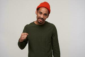 Portrait of young brown-eyed dark skinned brunette male with beard folding raised hand in fist and frowning his face while looking to camera, isolated over white background photo