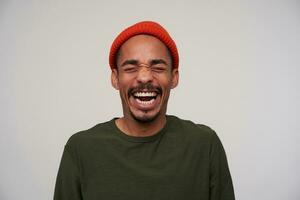 Good looking young joyful dark skinned brunette male keeping eyes closed while laughing cheerfully with wide mouth opened, standing against white background photo