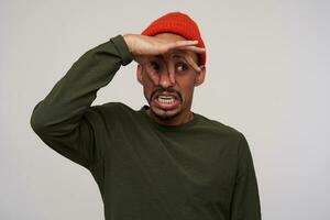 Studio photo of young pretty bearded dark skinned guy in casual clothes closing his nose and showing disgust while avoiding bad smell, standing against white background