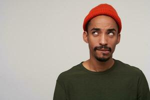 Puzzled young pretty dark skinned bearded brunette man biting underlip while looking upwards, plotting something, standing against white background in casual wear photo