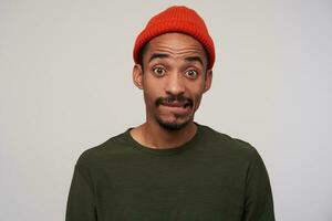 Bewildered young dark haired bearded man with dark skin wearing red hat while posing over white background, biting underlip and raising confusedly eyebrows while looking to camera photo