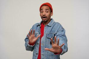 Fearful young brown-eyed dark skinned brunette man with beard protecting himself with raised hands and looking to camera with shocked face, isolated over white background photo