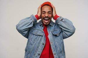 Stressed young good looking dark skinned brunette man with beard clutching his head with raised hands while standing over white background, frowning face while shouting emotionally photo