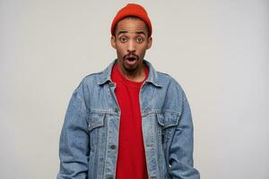 Dumbfounded young pretty dark haired bearded man with dark skin raising amazedly eyebrows while looking to camera with wide eyes and mouth opened, standing against white background photo