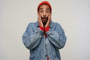 Shocked young handsome dark skinned brunette guy with beard holding face with palms and rounding eyes with raised eyebrows while standing over white background photo