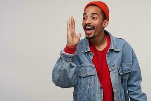 Indoor photo of cheerful lovely young bearded dark skinned guy dressed in red hat, red pullover and jeans smiling widely with raised palm while posing over white background