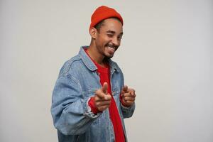 Indoor shot of young lovely dark skinned bearded brunette man winking at camera with cheerful smile, wearing red cap, pullover and blue jeans coat over white background with raised hands photo