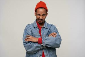 Studio photo of young dissatisfied bearded dark skinned brunette male frowning his face and folding hands while standing against white background in red hat, red pullover and jeans coat