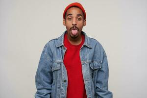 Portrait of young attractive brown-eyed dark skinned man with beard showing tongue to camera while posing over white background with hands down, dressed in casual wear photo