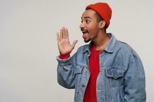 Side view of young attractive bearded dark skinned brunette hipster looking aside and screaming something, raising palm to his mouth while posing over white background photo