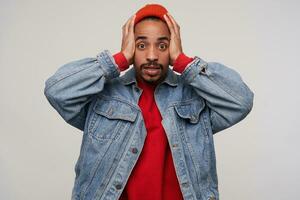 Bewildered young pretty dark haired bearded man with dark skin clutching his head with raised hands and looking perplexedly at camera, dressed in casual wear over white background photo