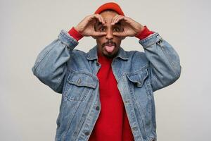 Funny shot of pretty young bearded dark skinned male showing tongue and making ridiculous faces while standing over white background in red hat, red pullover and jeans coat photo