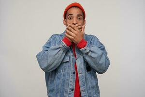 Photo of frightened young brown-eyed dark skinned bearded guy covering his mouth with raised palms while looking scaredly at camera, standing against white background