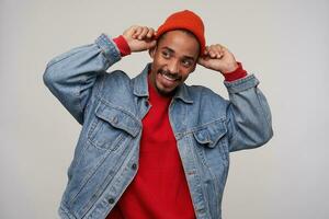 Pleased young lovely bearded brown haired man with dark skin raising hands to his headwear and looking cheerfully aside, wearing red cap, pullover and blue jeans coat over white background photo