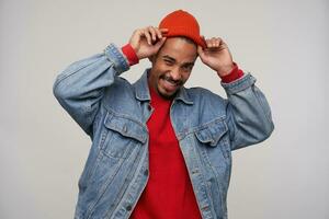 Cheerful young pretty dark skinned brunette male with beard holding his red cap with raised hands while standing over white background, being in nice mood and smiling happily photo