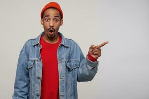 Amazed young handsome dark skinned bearded brunette male pointing aside with wide eyes and mouth opened, raising surprisedly eyebrows while posing over white background photo