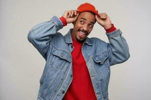 Studio photo of young charming dark skinned bearded guy looking happily aside with broad smile while keeping hands on his cap, isolated over white background in casual wear