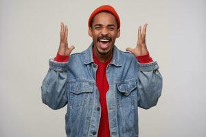 Excited young pretty dark skinned brunette male with beard frowning his face while screaming with wide mouth opened, raising emotionally hands while posing over white background photo