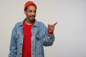 Photo of puzzled young dark skinned bearded guy frowning his eyebrows while looking aside with confused face, standing over white background with raised forefinger