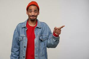 Studio photo of young dark skinned brunette man with beard getting air in his mouth and looking confusedly at camera, keeping index finger raised while posing over white background