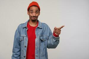 Confused young brown-eyed bearded dark haired male raising surprisedly eyebrows and puffing out cheeks while showing aside with index forefinger, isolated over white background photo