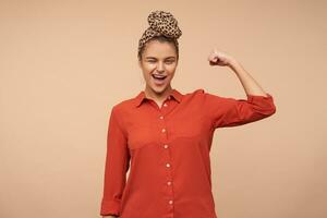 Cheerful young pretty brown haired lady with headband winking joyfully at camera while demonstrating her strong biceps with raised hand, isolated over beige background photo