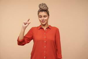Puzzled young brown haired lady wearing headband in knot while standing over beige background, looking confusedly on her hand while showing small size with fingers photo