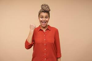 Excited young happy pretty brown haired woman with headband looking emotionally at camera while raising hand in yes gesture, isolated over beige background photo