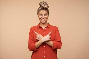 Happy young attractive brunette woman with headband smiling widely while pointing with forefingers in different directions, isolated over beige background photo