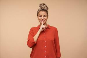 Positive young attractive brown haired lady with natural makeup keeping forefinger on her lips while asking to keep her secret and smiling gladly, isolated over beige background photo