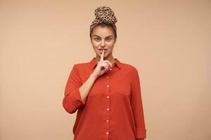 Indoor photo of young pretty brunette female raising hand in hush gesture while asking to keep silence, isolated over beige background in headband and red shirt