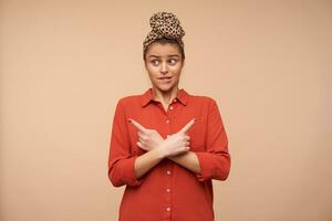 Bewildered young pretty brunette woman with headband biting worringly underlip while showing in different directions with index fingers, posing over beige background photo