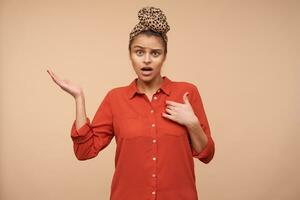 Bewildered young pretty brunette lady with natural makeup showing on herself and keeping palm raised while looking confusedly at camera, standing over beige background photo