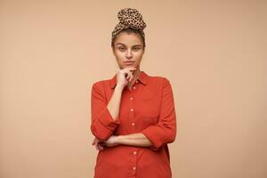 Pensive young pretty brunette lady squinting her eyes while looking thoughtfully at camera and holding chin with raised hand, isolated over beige background photo