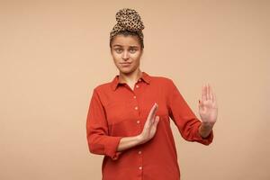 Indoor photo of young pretty green-eyed brunette lady keeping her lips folded while looking at camera and raising her palms in refusing gesture, isolated over beige background