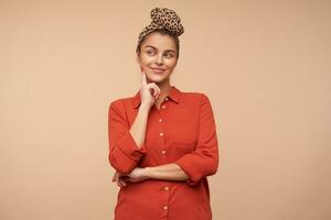 Positive young lovely brunette woman with natural makeup smiling pleasantly while looking dreamily aside and keeping forefinger on her cheek, standing over beige background photo