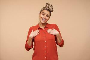 Pleased young pretty brunette woman wearing headband in knot while posing over beige background, being in nice mood and smiling gladly at camera while keeping her hands on chest photo