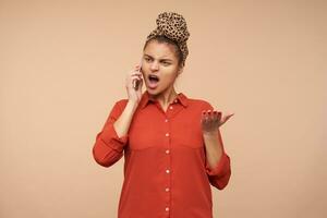 Angry young green-eyed brunette female with headband raising emotionally hand and shouting crossly while having unpleasant phone talk, standing over beige background photo