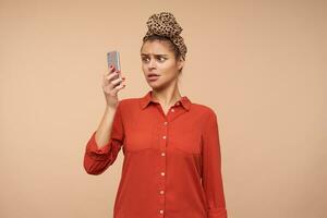 Indoor photo of confused young brunette woman wearing headband in knot while posing over beige background, keeping mobile phone in raised hand and looking confusedly on screen