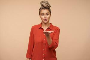 Indoor photo of young attractive brown haired female folding her lips in air kiss and looking positively at camera, standing over beige background with raised hand