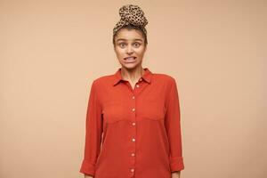 Studio shot of bewildered young brown haired lady twisting her mouth while looking surprisedly at camera, posing over beige background in casual clothes photo