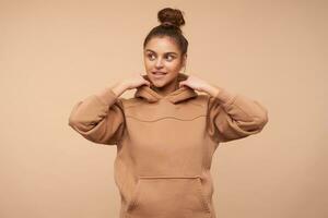 Positive young attractive female wearing her brown hair in knot while standing over beige background, looking aside with light smile and raising hands to her hood photo