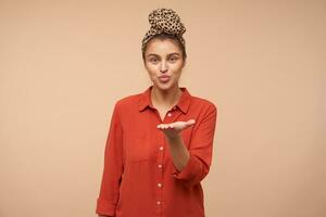 Pleased young attractive green-eyed brunette female pursing her lips while sending air kiss at camera, standing over beige background with raised hand photo