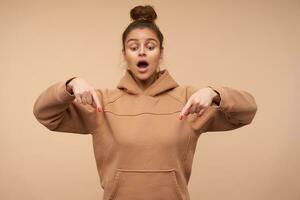 Agitated young pretty brown haired woman in sporty clothes keeping her hands raised while pointing down with index fingers, standing over beige background photo