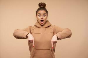 Overjoyed young pretty brown haired female looking at camera with wide eyes opened while showing down, standing over beige background in nude sweatshirt photo