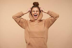 Good looking young pretty brunette woman keeping her eyes closed while laughing happily, raising hand to her hood while standing against beige background photo