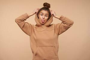 Funny photo of young lovely brown haired lady with bun hairstyle showing cheerfully tongue at camera while taking on her hood, isolated over beige background